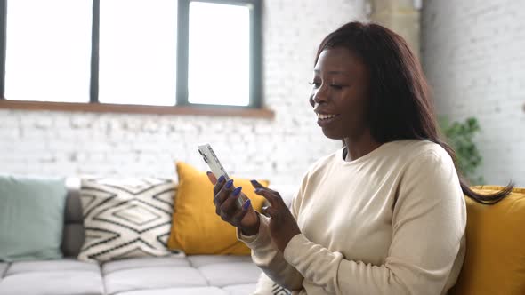 Inspired Smiling Multiracial Woman Using Smartphone Sitting Down on Comfortable Couch