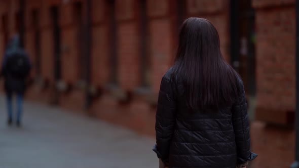 a Woman with Long Hair and a Dark Coat Smiles and Walks Against the Background of a Brick Building alt