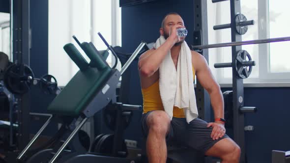 Strong Man Bodybuilder Sitting on Bench Resting and Drinking Water in Gym