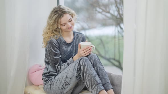Charming Happy Smiling Woman Sitting on Windowsill Holding Coffee Cup Looking Out the Window alt