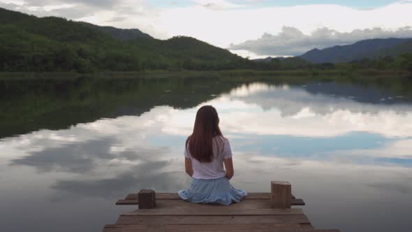 Lonely young woman sitting on wooden bridge and looking at beautiful landscape at sunset alt