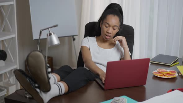 Exhausted Overburdened Young Woman with Feet on Table Yawning Surfing Internet on Laptop in Home alt