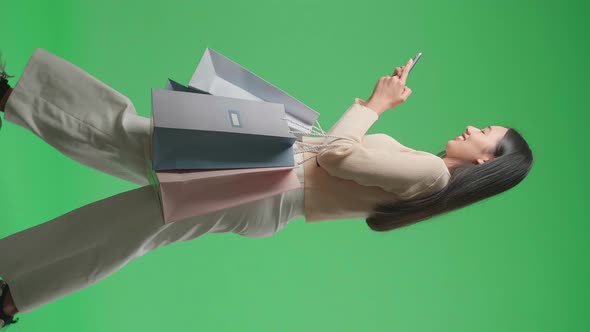 Side View Of Woman With Shopping Bags Using Mobile Phone While Walking In Front Of Green Screen alt