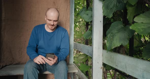 Man is Sitting on the Porch of a Village House and Reading an Electronic