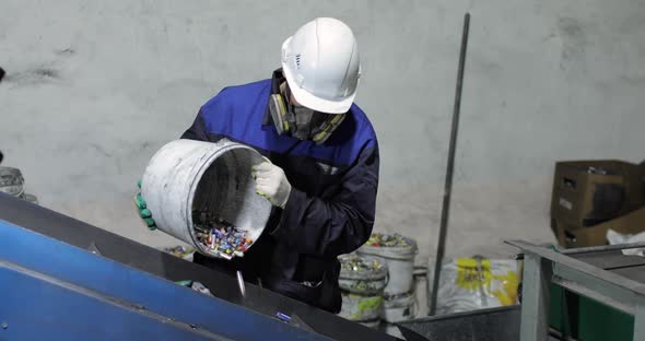 A Man in Uniform Pours Used Batteries From Bucket Onto a Moving Conveyor Belt alt