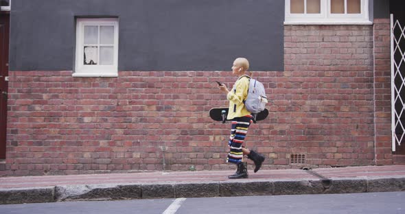 Mixed race woman walking on street holding her skateboard and her phone alt
