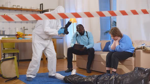 Forensic Specialist Taking Photo of Dead Body While Policeman Interrogating Witness Sitting on Couch alt