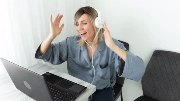 Attractive european blonde woman in headphones listening music and dancing sitting on comfy chair