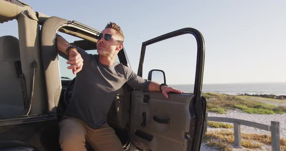 Happy caucasian man in sunglasses sitting in car with open door on sunny day at the beach alt