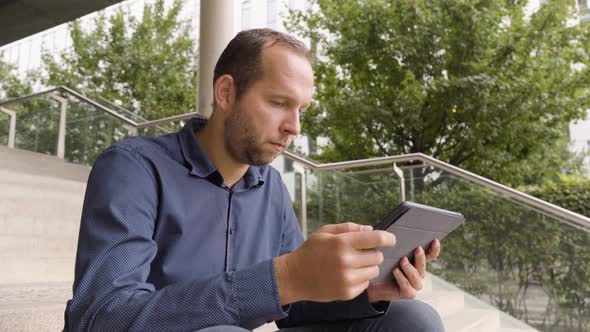 A Caucasian Man Looks at a Tablet As He Sits on a Staircase in an Urban Area  Closeup From Below