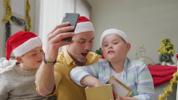 Caucasian father and two sons opening gifts while having a videocall on smartphone during christmas alt