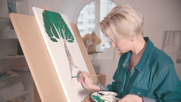 A Young Blonde Woman Draws a Tree with Paints in the Art Studio alt