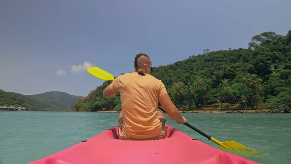 Man with Sunglasses and Hat Rows Pink Plastic Canoe Along Sea Against Green Hilly Islands with Wild