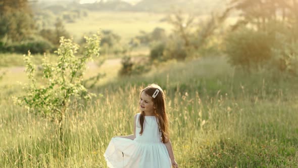Little Cute Girl with Flowing Hair in a White Summer Dress is on a Green Lawn Laughing and Posing in