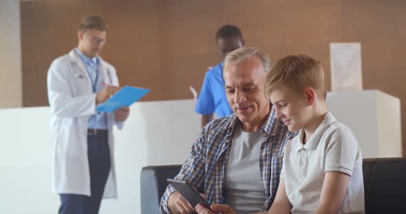 Father and Son Using Digital Tablet Sitting in Hospital Waiting Room alt