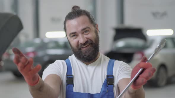 Portrait of Successful Mechanic Makes Inviting Gestures and Smiles Holding Huge Wrench in Hands alt