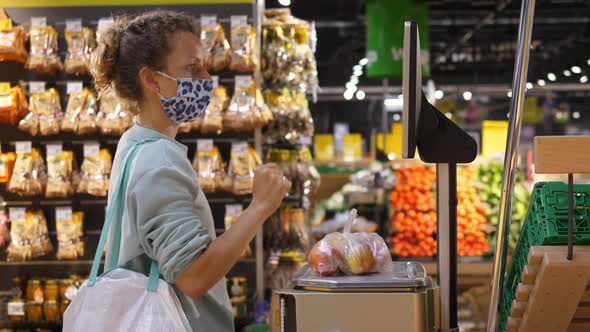 Side View of a Blonde Girl in a Face Mask Weighing Fruits on an Electronic Scale in a Supermarket alt