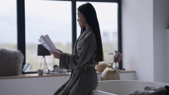 Young Slim Asian Woman Looking Through Business Documents Sitting on Bathtub in Bathrobe alt