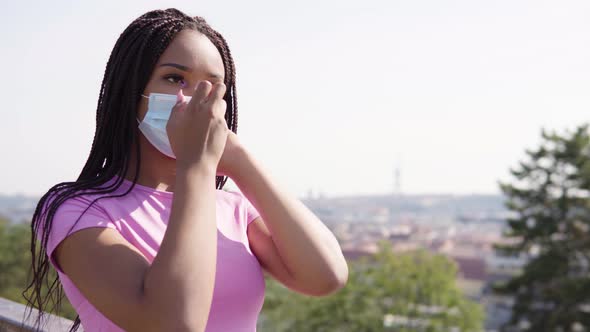 A Young Black Woman Puts on a Face Mask and Looks at the Camera - a Cityscape in the Background alt