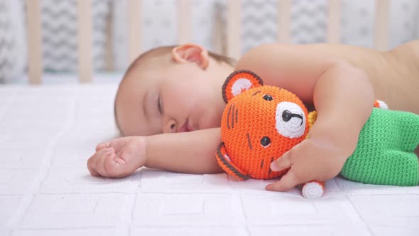 Toddler 12-17 months old sleeps in bed hugging a knitted tiger toy. Selective focus. alt