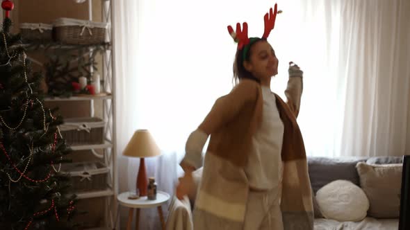 Young Cheerful Pretty Woman Wearing Reindeer Antlers Headband Dancing with Christmas Lights at Cozy alt