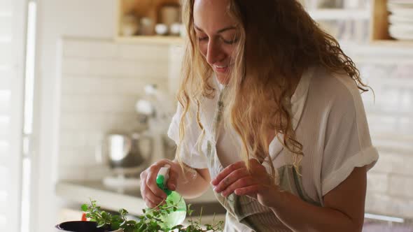 Smiling caucasian woman watering potted plants standing in cottage kitchen alt