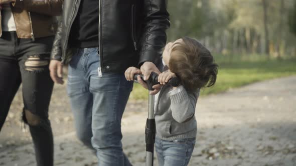Cheerful Cute Caucasian Boy Riding Scooter in Sunny Park with Unrecognizable Parents alt