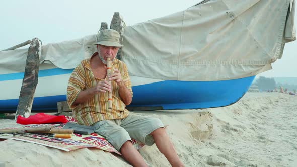 Senior man playing bamboo flute on the beach next to fishing boat alt