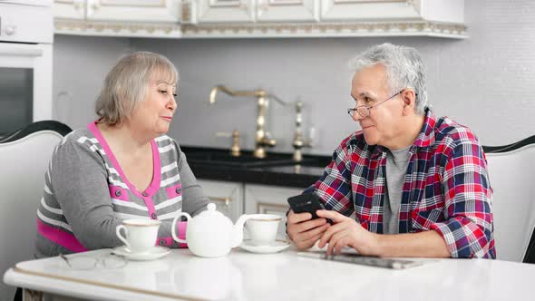 Aged Couple Smiling Talking Using Smartphone During Breakfast at Kitchen alt