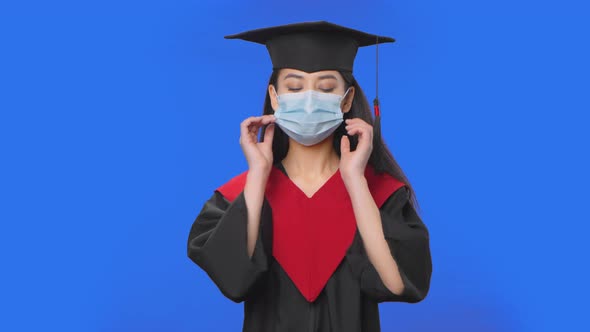 Portrait of Female Student in Cap and Gown Graduation Costume Removes Protective Medical Mask alt