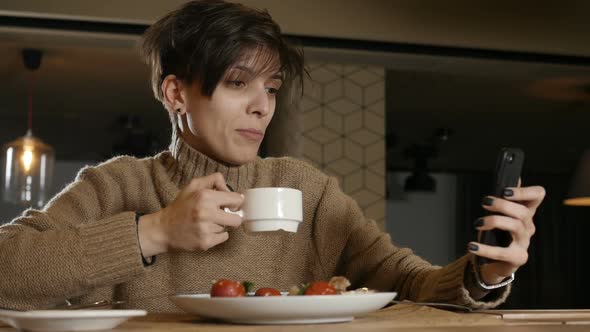 a Woman with a Short Haircut Looks at the Phone and Drinks Coffee