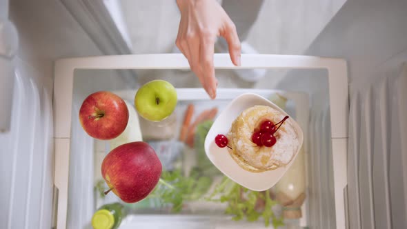 Woman's hands choosing between apples and cake from refrigerator.