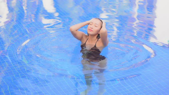 High angle. An attractive young woman in a swimming pool smooths back her wet hair then looks around alt