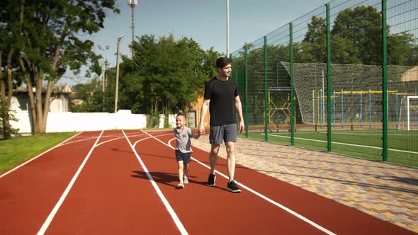Dad and Child Boy in Sportswear are Smiling Holding Hands and Walking at the Stadium alt