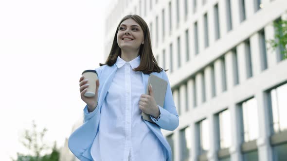 Woman With Tablet and Coffee Cup Walking Between Business Buildings and Smiling With Joy