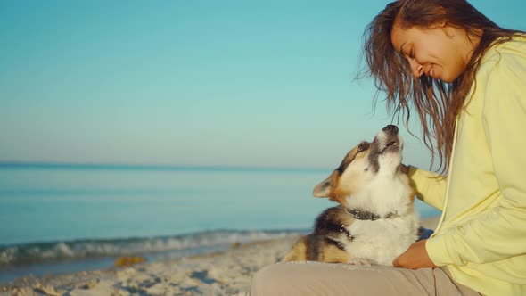 Candid Laughing Latin Girl in Yellow Hoodie at Summer Beach with Cute Pet Corgi Dog Sitting in Front alt