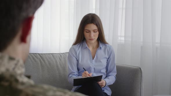 Young Woman Psychologist Filling Anamnesis for Man Soldier Asking Patient and Writing in Notepad at alt
