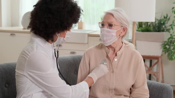 Afro American Woman Doctor in Mask Listens to the Breathing of an Elderly Woman Sitting on the Couch alt