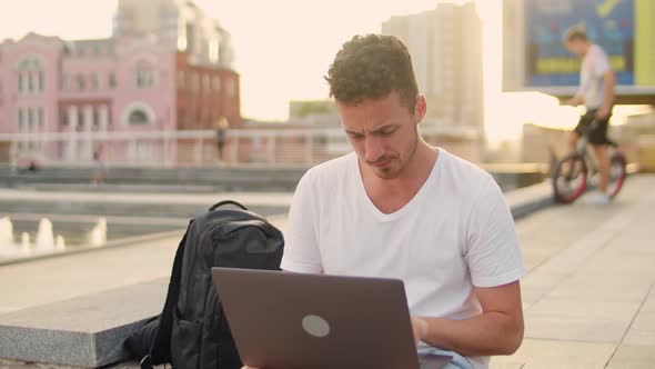 Young Handsome Man in White Tshirt Works with Laptop in the City Center While Sitting on the Steps alt