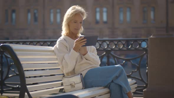 Senior Woman with Tablet Pc Computer Sitting on City Bench By Riverside alt