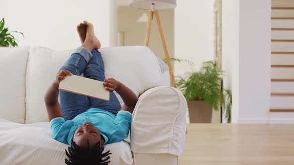 African american boy smiling while using digital tablet while lying on the couch at home alt
