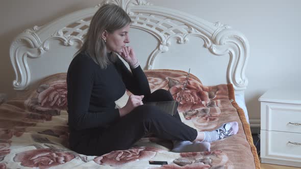 A Middleaged Woman Sits on Her Bed and Holds a Laptop in Her Arms alt