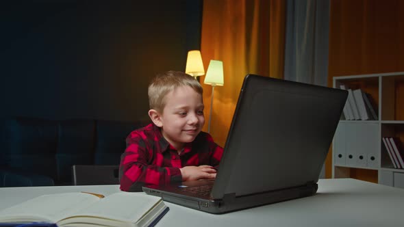 The Child Teaches Lessons While at Home Sitting in the Living Room at the Table Using a Laptop Books alt