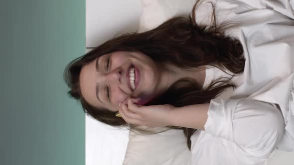 Vertical Shot Happy Young Woman in White Pajamas Talking on Mobile Phone in Bed and Smiling