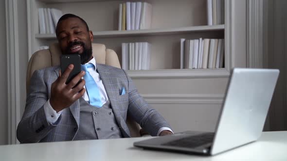Black Man Is Sitting in Armchair in His Office and Talking By Video Call in Modern Phone alt