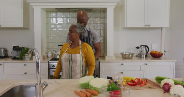 African american senior man tying apron from back to his wife in the kitchen at home alt