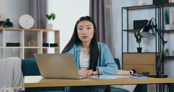 Asian Girl with Long Black Hair Sitting at Her Workplace in Home Office and Looking Into Camera alt