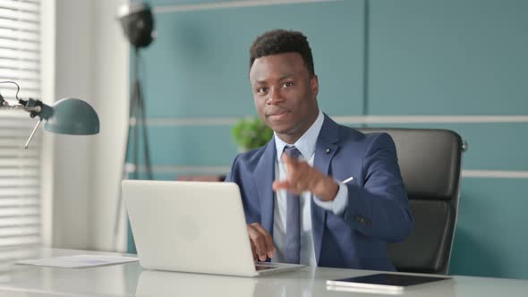 African Businessman Pointing at Camera While Using Laptop in Office alt
