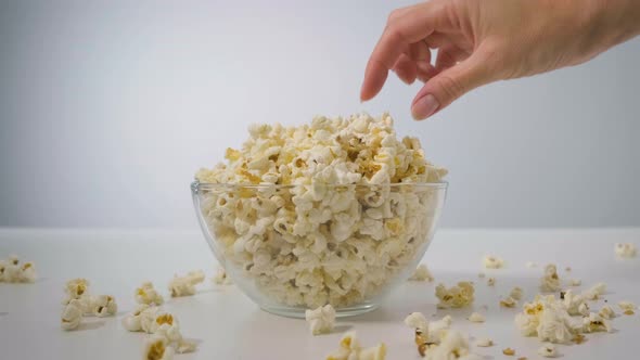 Closeup Woman's Hand Takes Popcorn From Transparent Bowl on White Table Against Background of Light alt