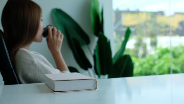 A young asian woman grabbing a coffee cup to drink and relaxing after reading book at home